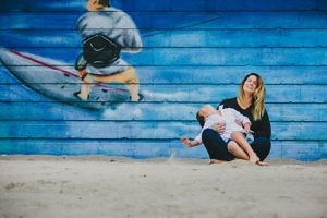 son-sitting-on-mothers-lap-on-a-sandy-beach-laugh-hysterically-in-front-of-a-windsurfing-mural