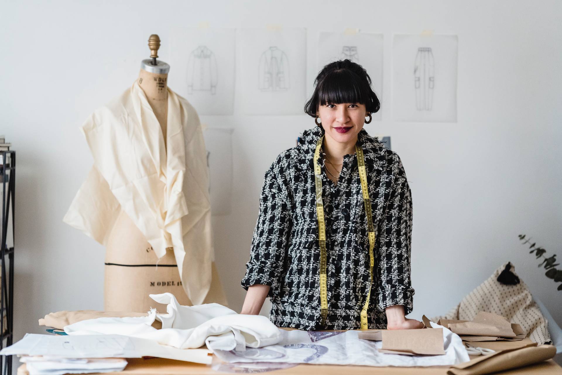 a fashion designer and creator stands in her studio wearing a black and white plaid shirt. A measuring tape around her neck standing in front of a mannequin with a half finished project