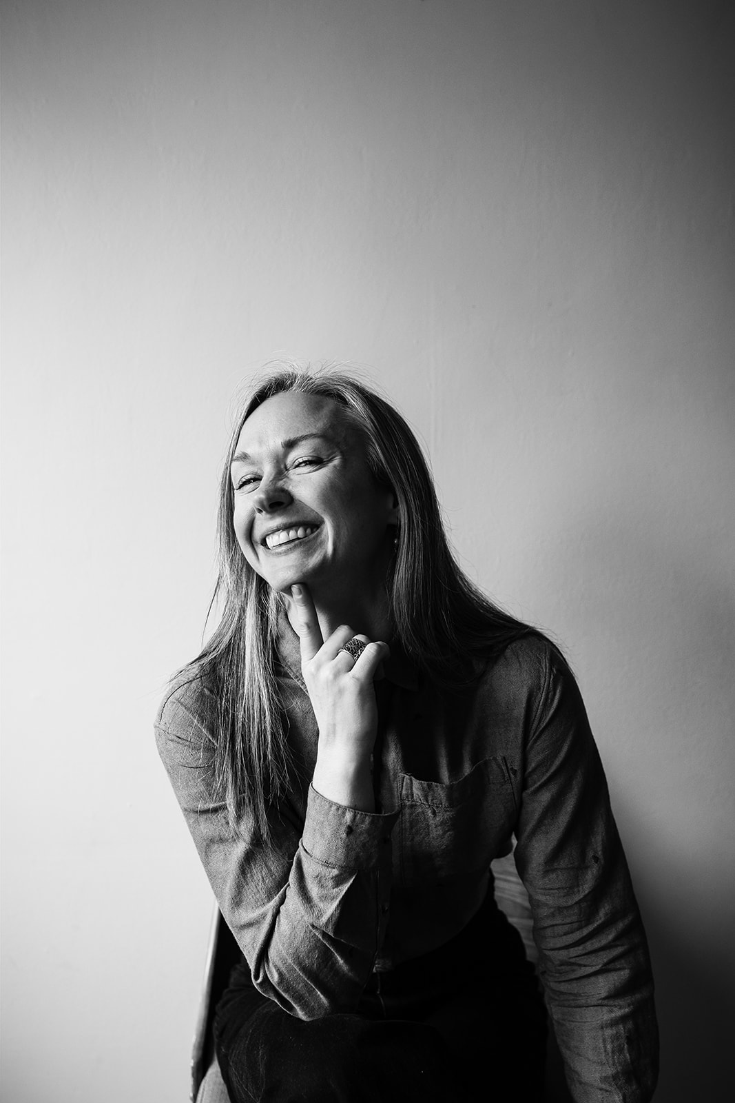 black and white photographs of sex educator business headshot. sitting on stool smiling at the camera with long grey hair and hand under chin. 