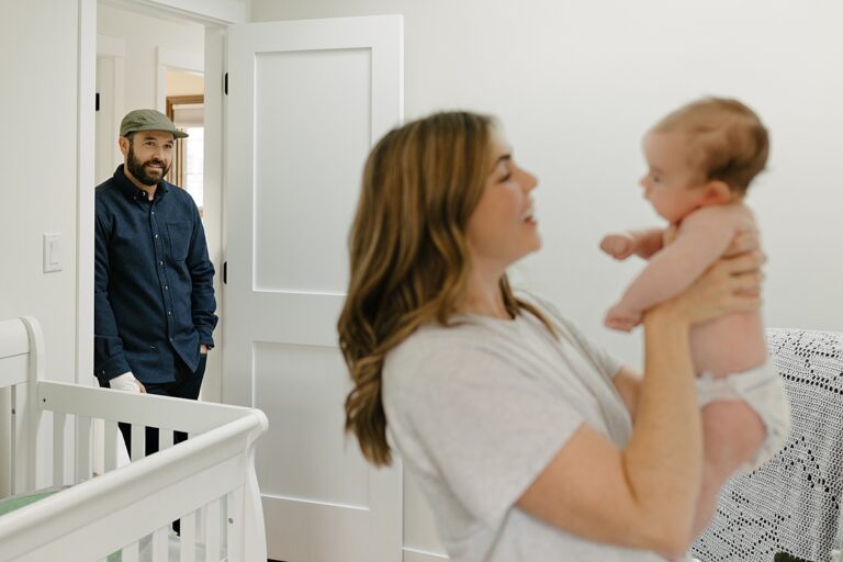a father watches happily from the doorway of his sons nursery his wife lift his newborn son out of his crib